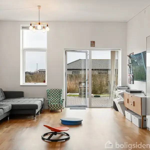 Grå sofa står i en lys stue med trægulve. Glasdøre og vinduer viser en have udenfor. Fjernsyn på væggen, hylder med bøger og legetøj på gulvet.