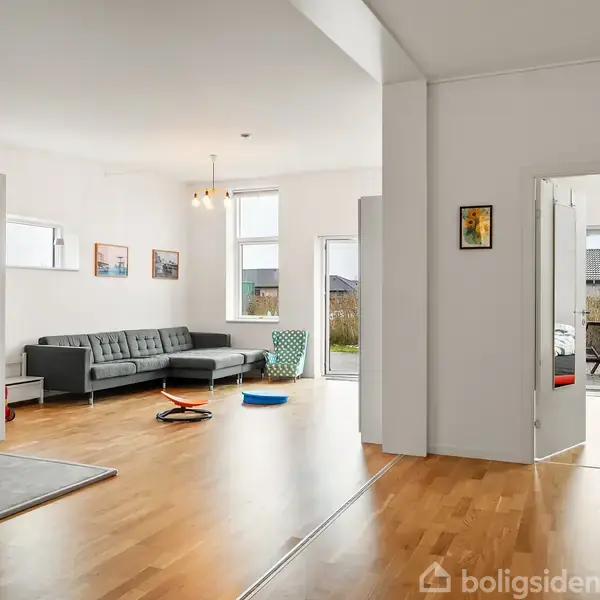 Gråt sofaarrangement står i en stue med trægulv, to billeder på væggen og store vinduer med udsigt til en have. En åben dør leder til et andet rum.