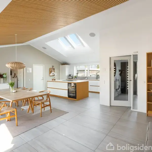 Spisestue med langbord omgivet af træstole, stående på lyst gulvtæppe. I baggrunden findes et moderne køkken med hvide skabe og ovenlysvinduer. En åben hylde står på gulvet.