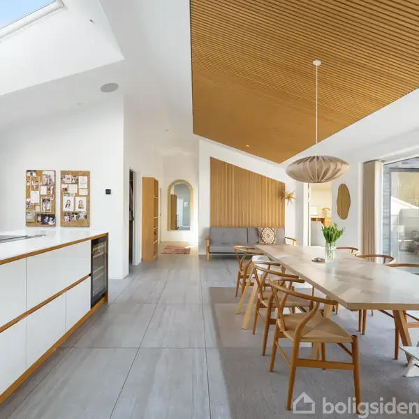 Et stort, lyst køkken-alrum med spisebord og stole i træ. Ovenlysvindue og glasparti til terrasse giver naturligt lysindfald. Moderne indretning med træloft og hvide vægge. Ingen tekst i billedet.