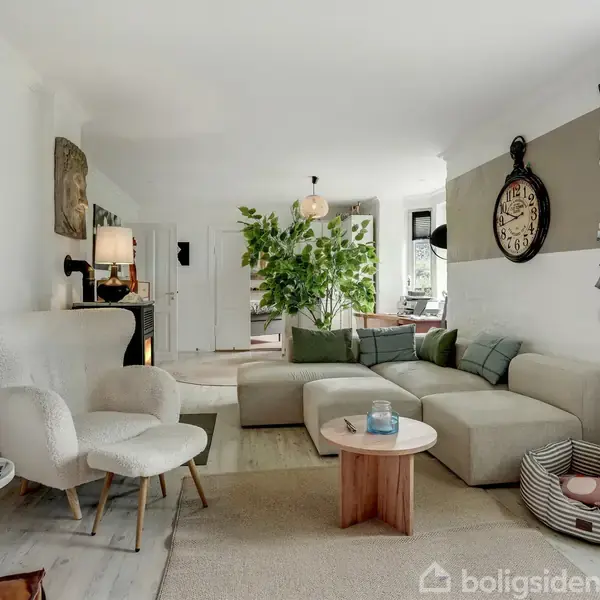 Stue med lys indretning, indeholder en beige sofa, lænestol, runde borde, og hængende ur på væggen. En plante i baggrunden og hundekurv ved siden af radiatoren.