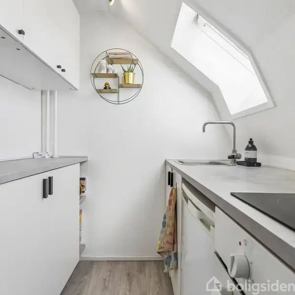 Et lille, lyst køkken med hvidt interiør. På bordpladen står en blå tekande, chokoladepulver og elkedel. Et rundt væghylde indeholder planter samt en gul krukke. En skråvæg med ovenlys.