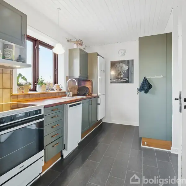 Et køkken med en sort komfur, grå skabe og hvidevarer. En plante ses på vindueskarmen, og en viskestykke hænger på et skab. Væggene er lyse.