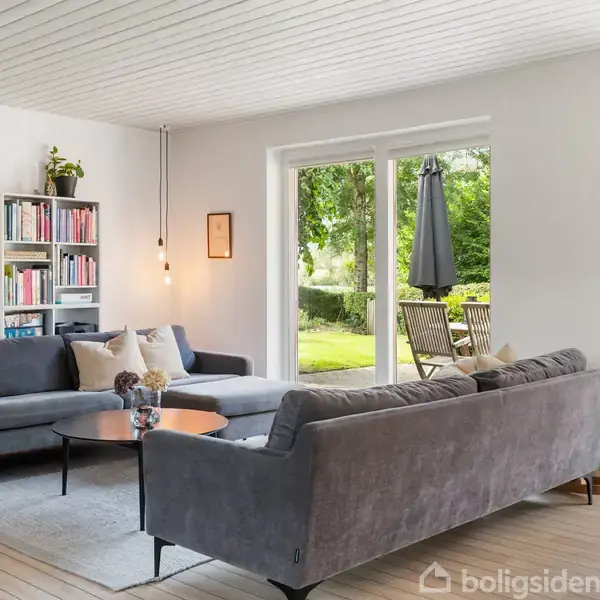 To store grå sofaer står i vinkel omkring et rundt bord, omgivet af en bogreol og en dør til en terrasse med grøn udsigt. En hængelampe lyser rummet.