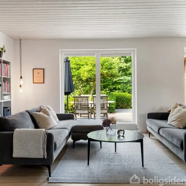 Grå sofaer står overfor hinanden i en stue med træloft. En glasdør fører ud til en terrasse med havemøbler, omgivet af grønne træer. En reol står til venstre.