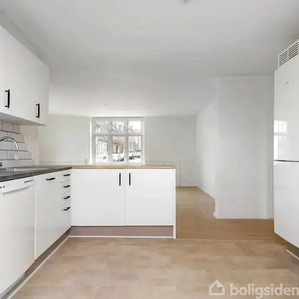 Hvidt køkken med skabslåger og skuffer, overskabe, indbygget ovn og komfur til højre. Fliser på væggen ved vasken. Rummet har et vindue i baggrunden.