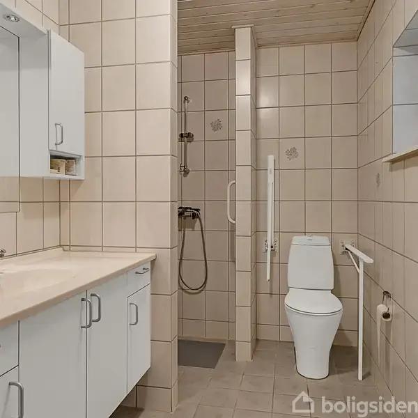 Badeværelse med beige fliser. Håndvask og spejlskab til venstre, bruser i midten, og toilet til højre. Rummets indretning er funktionel og moderne.