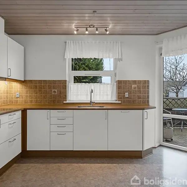 Et køkken med hvide skabe og brun bordplade står i hjørnet. En vask og vandhane er under et vindue med hvidt gardin. En dør fører ud til en træterrasse.