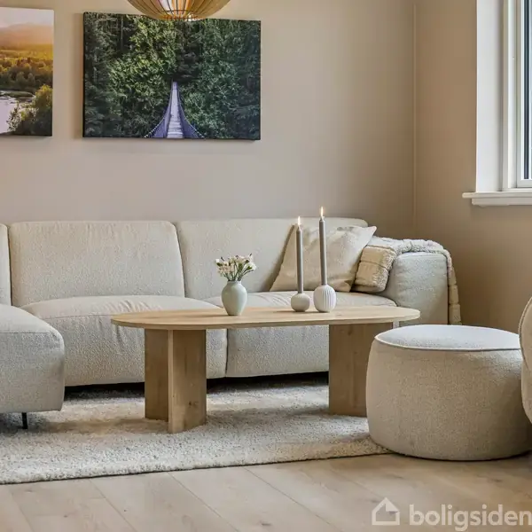 En beige sofa står i en hyggelig stue, flankeret af et lavt træbord med dekorative lys og blomster. På væggen hænger naturbilleder, mens et vindue tilføjer lys.