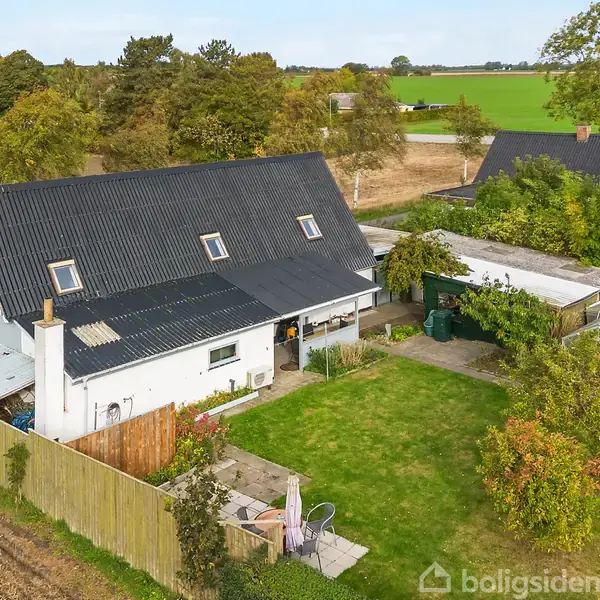 Et hvidt hus med sort tag står i en grøn have, omgivet af buske og træer. Der er en terrasse med havemøbler og en træplankehegn.
