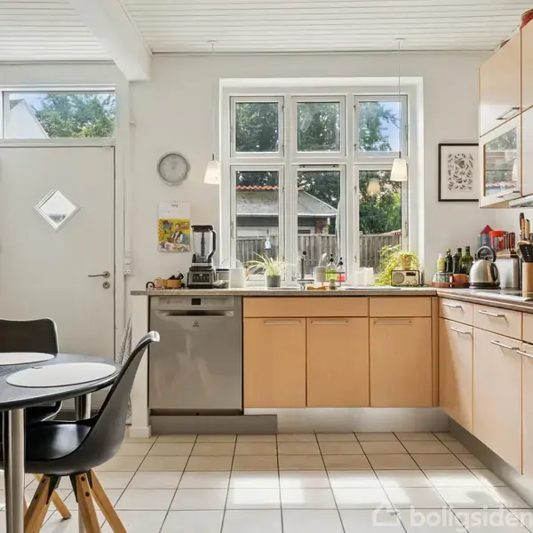 Et køkken med lyse træskabe og hvide vægge har en sort spisebord med fire stole. Et vindue med udsigt til en have bringer naturligt lys ind.