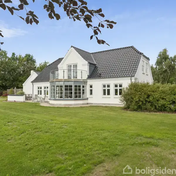 Hvidt hus står stille omgivet af en stor grøn græsplæne og træer. Taget er mørkegråt, og der er en balkon på første sal.