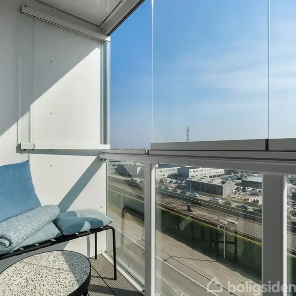 En lille balkon med en pude og tæppe på en stol, ved siden af et lille bord. Udsigt til en vej og industrielle bygninger under en klar himmel.