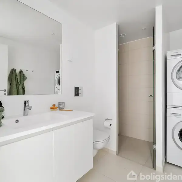 Vaskesøjle med vaskemaskine og tørretumbler står i et hvidt badeværelse. Ved siden af er der en toiletstol og en lang håndvask med spejl. Grønne håndklæder ligger på vasken.