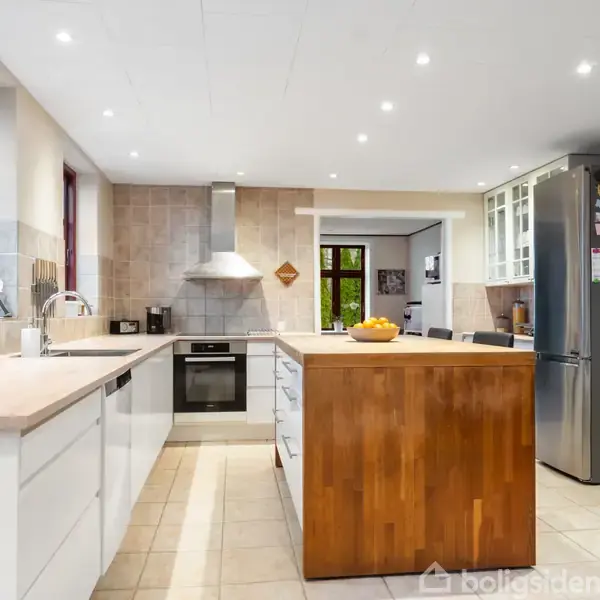 Et køkken med en central ø viser en frugtskål. Rummet er lyst og moderne med hvide skabe, fliser på væggene, og indbyggede apparater. En grå køleskab står til højre.