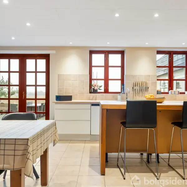 Et moderne køkken med et spisebord dækket af en ternet dug står til venstre. To høje stole ved en køkkenø i midten. Rødbrune vinduesrammer slipper lys ind.