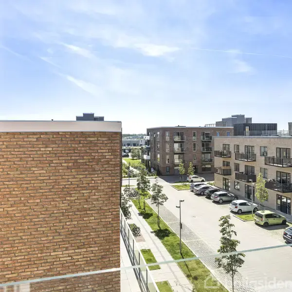 En række moderne lejlighedsbygninger står på en solrig dag, omgivet af parkerede biler og velplejede grønne områder, alle set fra en højere etages synsvinkel.