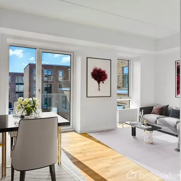 En lys stue med en grå sofa og sofabord står i baggrunden. Et spisebord med en stol og blomster står nær vinduet, som åbner ud mod en altan. Kunstværker pryder de hvide vægge.