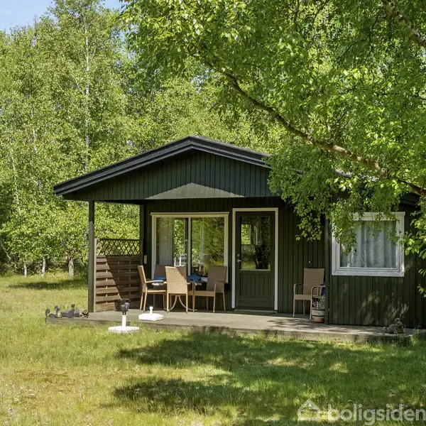 Sommerhus i træ står stille på en grøn græsplæne omgivet af træer. Terrassen har havemøbler, og huset er malet mørkt med lyse detaljer. Atmosfæren er fredfyldt og naturlig.
