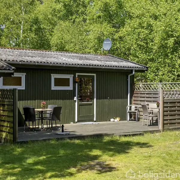 Et træhus med en mindre terrasse har borde og stole placeret udenfor. Omgivet af frodige grønne træer og en græsplæne i et roligt, landligt område.