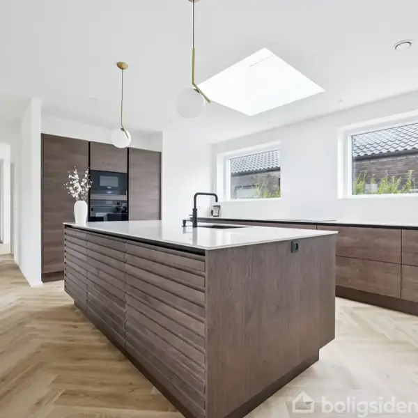 Køkkenø med mørkt træ og vask står i et lyst rum med parketgulv. Store vinduer tillader naturligt lys, mens pendellamper hænger ovenover. Skydedøre fører til andre rum.