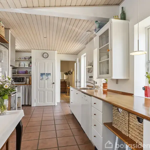 Et lyst køkken med hvide skabe har en træbordplade. En vase med blomster står på spisebordet til venstre. Vinduer og planter til højre. En dør og vægur ses i baggrunden.