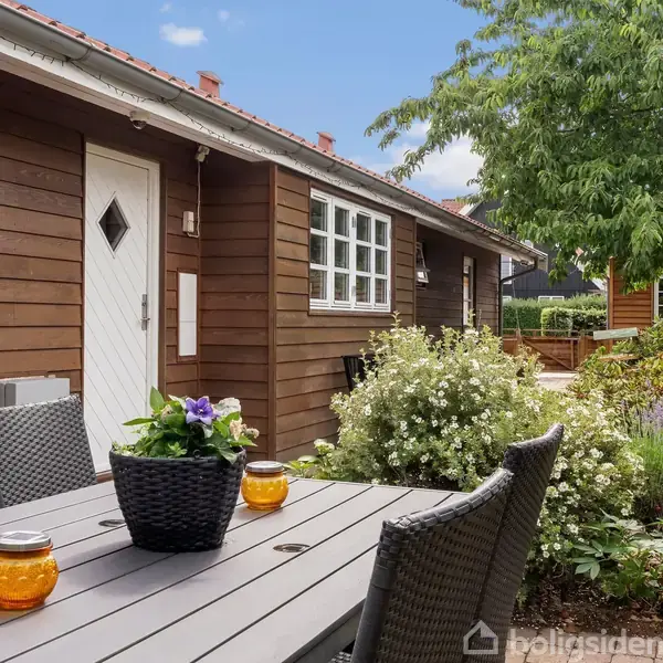 En havebord med flettede stole står på en terrasse. Bordet har en sort blomsterkrukke og to orange lanterner. I baggrunden ses et træhus omgivet af buske og træer.