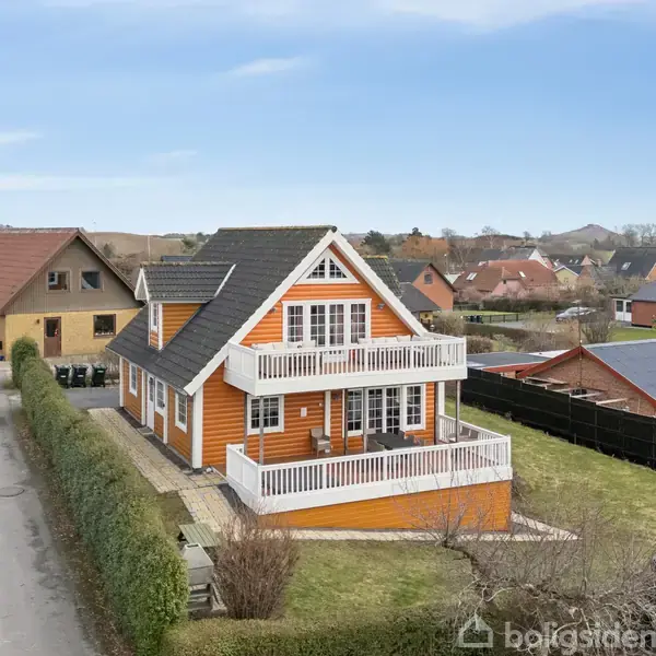 Et to-etagers træhus med hvid balkon står på en villavej omgivet af hække og andre huse i et forstadskvarter. Skarpt afgrænset have ses bag huset.