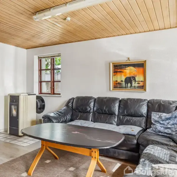 Sort lædersofa står i stue ved siden af sofabord og under billede af elefanter. Træloft og vinduer giver et hyggeligt, lyst rum. Køkken ses i baggrunden.