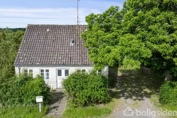 Et hvidt hus står stille omgivet af grønne buske og et stort træ. En lille sti fører til hoveddøren. Husk at ignorere vandmærket "Boligsiden".