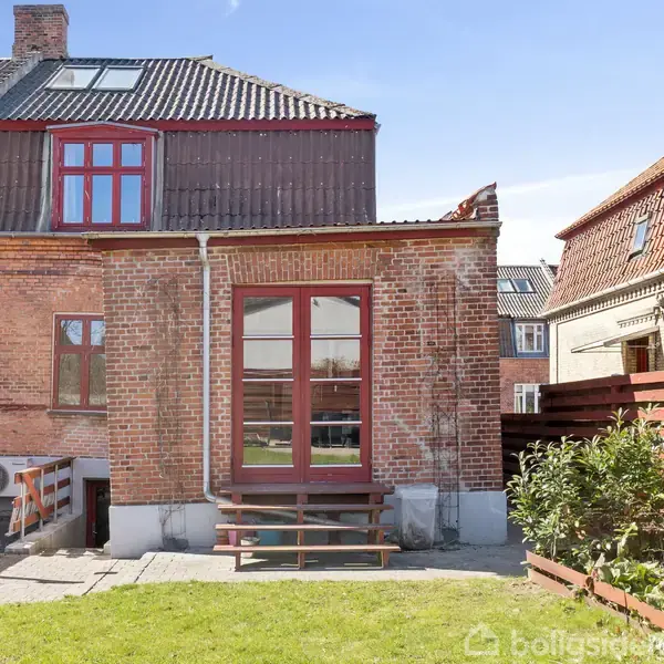 Rødstenshus med store vinduer står i solrigt, åbent haveområde. Kort trappe leder fra bagdør til grøn græsplæne. Naboens hus og et hegn ses til højre.