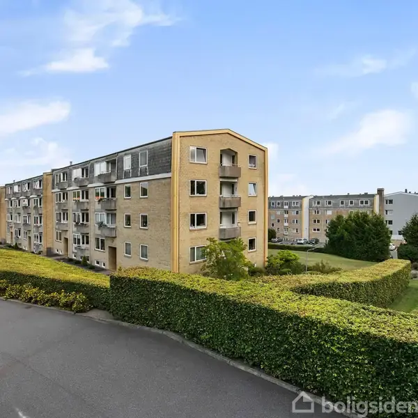 Et lejlighedsbyggeri står langs en vej, omkranset af pæne hække. Bygningen er lavet af mursten og har flere etager. I baggrunden ses flere lignende bygninger samt græsarealer.