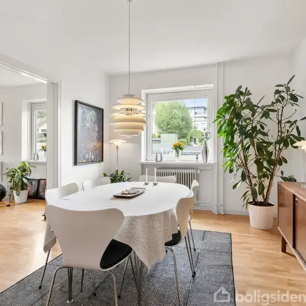 Et spisebord med hvide stole står i et lyst rum. Over bordet hænger en designerlampe. Der er store grønne planter og en brun kommode nær vinduet med udsigt til træer udenfor.