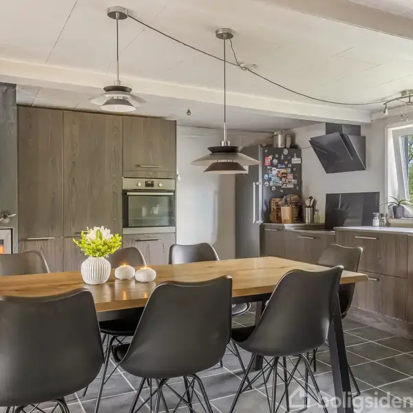 Køkken med spisebord og sorte stole, pejs til venstre og moderne grå køkkenelementer med rustfri apparater. Store vinduer slipper naturligt lys ind. Blomster er placeret på bordet.