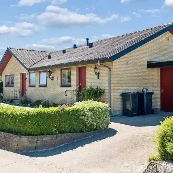 Gulstensvilla med sort tag står stille i en forstadshave omkranset af en lav hæk. Skraldespande er placeret foran en garage med røde døre.