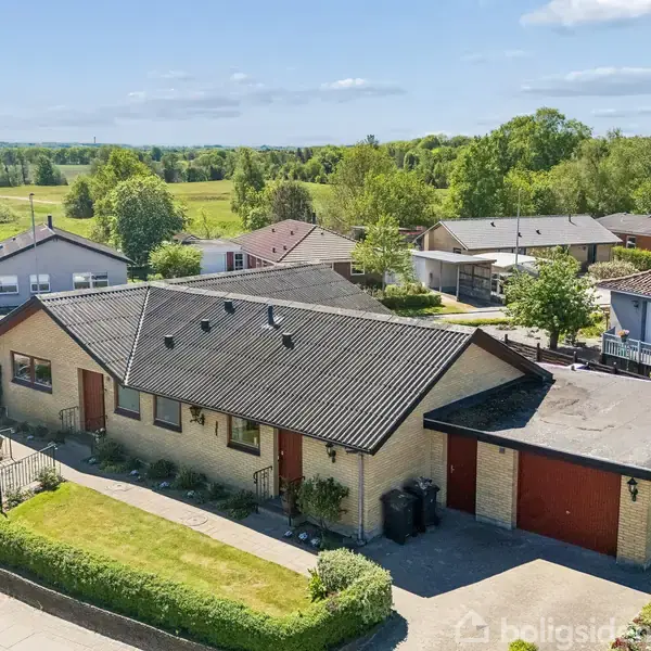En en-etagers murstensvilla med et sort tag, placerer sig på en velholdt græsplæne med indkørsel og garage. Omgivet af flere huse i et naturskønt område med marker i baggrunden.