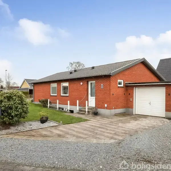 Rødt murstenshus med garage, står omgivet af en grusindkørsel og grøn forhave i et parcelhuskvarter.