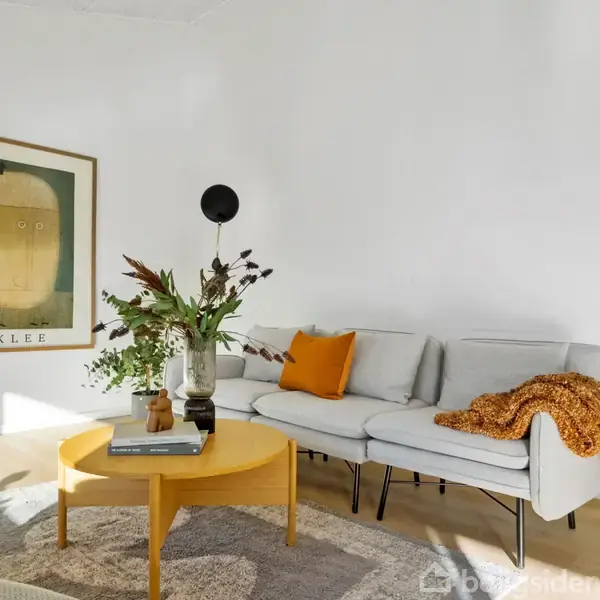En lysegrå sofa er pyntet med en gul pude og et orange tæppe. Foran sofaen står et rundt, gult sofabord med en vase med blomster. En vægplakat med teksten "KLEE" hænger ved siden af.