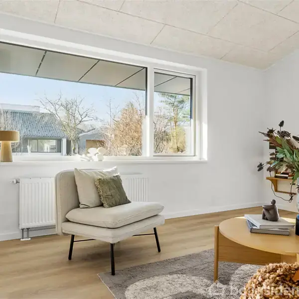 Stol står ved siden af et stort vindue, og en pude ligger på sædet. Rundt sofabord med bog og vase i en lys stue med trægulv og udsigt til have.