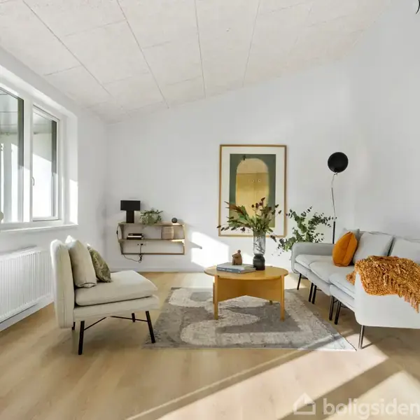 Stue med sofa og lænestol, der omkranser et sofabord. En stor vindueskarm til venstre lader lys ind. Vægdekoration og planter giver rummet liv.