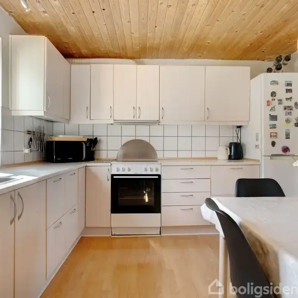 Hvidt køkken med komfur og ovn i midten, omgivet af skabe. Trægulv og loft. Et køleskab med magneter står til højre og et bord med blomster til venstre.