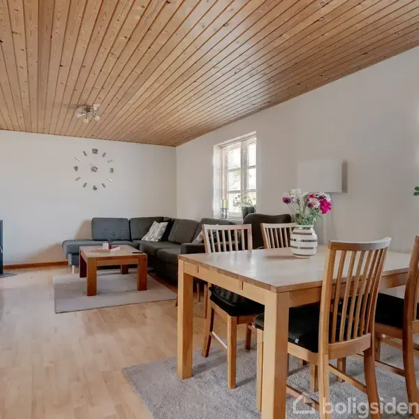 Stue med træloft og lyst trægulv, hvor en grå hjørnesofa står ved vinduer. Et spisebord med træstole står til højre. Pejs i hjørnet, ur på væggen.