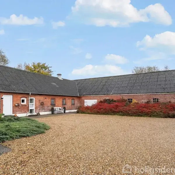 En lang, brun murstensbygning med mørkt tag står stille i et åbent gårdmiljø omgivet af grus og grønne buske under en blå himmel med skyer.