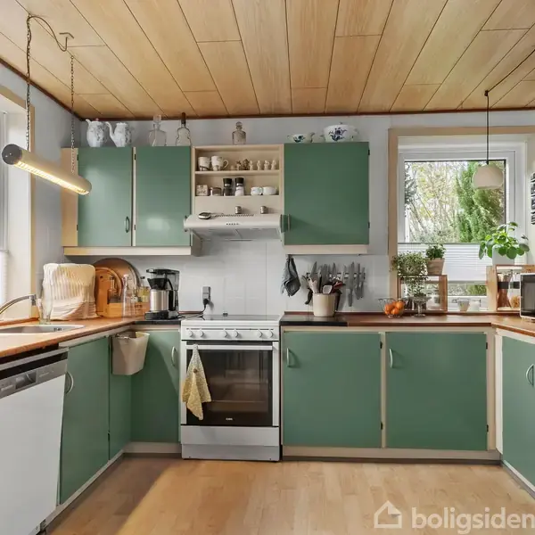 Køkken med grøntskabslåger og træbordplader, der indeholder en komfur med emhætte, vask, opvaskemaskine og køleskab. Køkkenet er lyst op af vinduer og har planter og køkkenredskaber som dekoration.