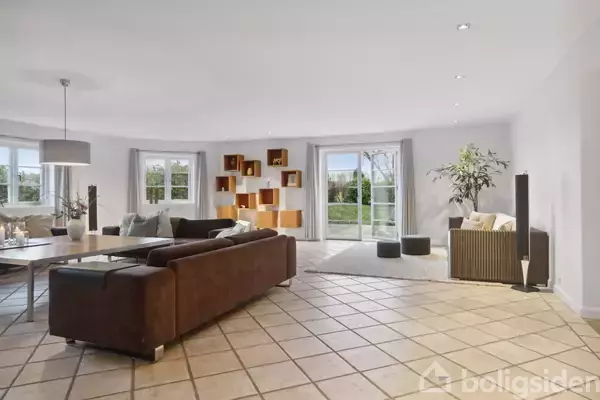 Brun sofa står i moderne, lyst stueområde med fliset gulv. Store vinduer og glasdøre åbner op til en grøn have. Hylder og planter tilføjer dekoration.