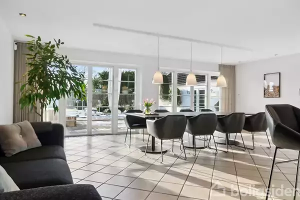 Stort spisebord omgivet af centrale lamper og stole, placeret i en lys stue med flisegulv, store vinduer og en frodig plante ved siden af en mørk sofa.