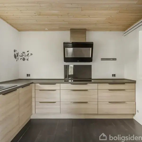 Køkken med lyse træskabe og sort bordplade; indbygget ovn og emhætte på væggen. Trægulv og loft tilføjer naturmaterialer, mens et lille vindue giver naturligt lys. Ingen tekst fundet.