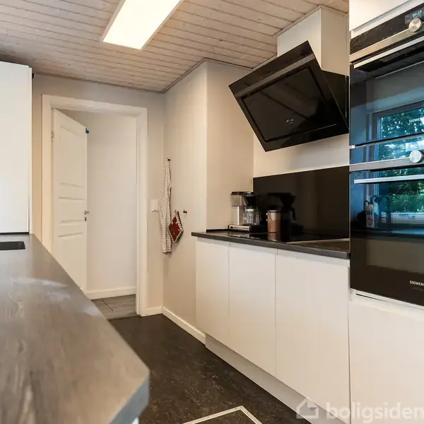 Et moderne køkken med hvide skabe og sorte apparater såsom en ovn og emhætte. Der er en køkkenbordplade langs væggen, og et vindue til højre giver udsigt til det grønne udenfor.