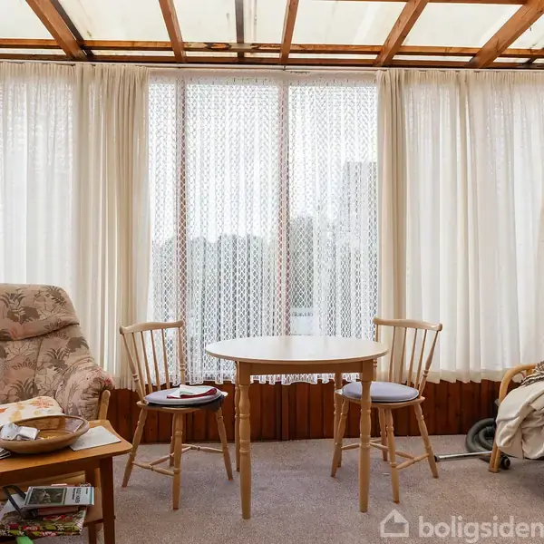 Et møbleret rum med en polstret lænestol til venstre og to træstole omkring et rundt bord. Gardiner i baggrunden, tæppe på gulvet, afslappet atmosfære.