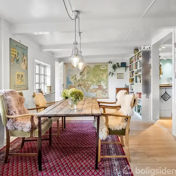 Et træbord omgivet af polstrede stole er placeret på et rødt tæppe i en lys spisestue. Vinduer og vægdekorationer skaber en hyggelig atmosfære med hylder langs væggen.